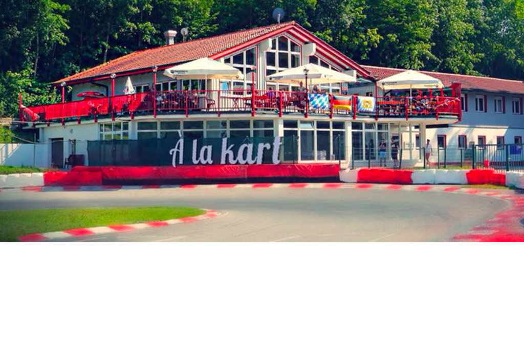Gokart-Bahn mit einer Kurve im Vordergrund, gesäumt von roten und weißen Barrieren. Im Hintergrund ist ein großes Gebäude mit einer Terrasse mit der Aufschrift „A la kart“ zu sehen, mit Sonnenschirmen und einem Restaurant-Setup. Bäume umgeben die Szene.