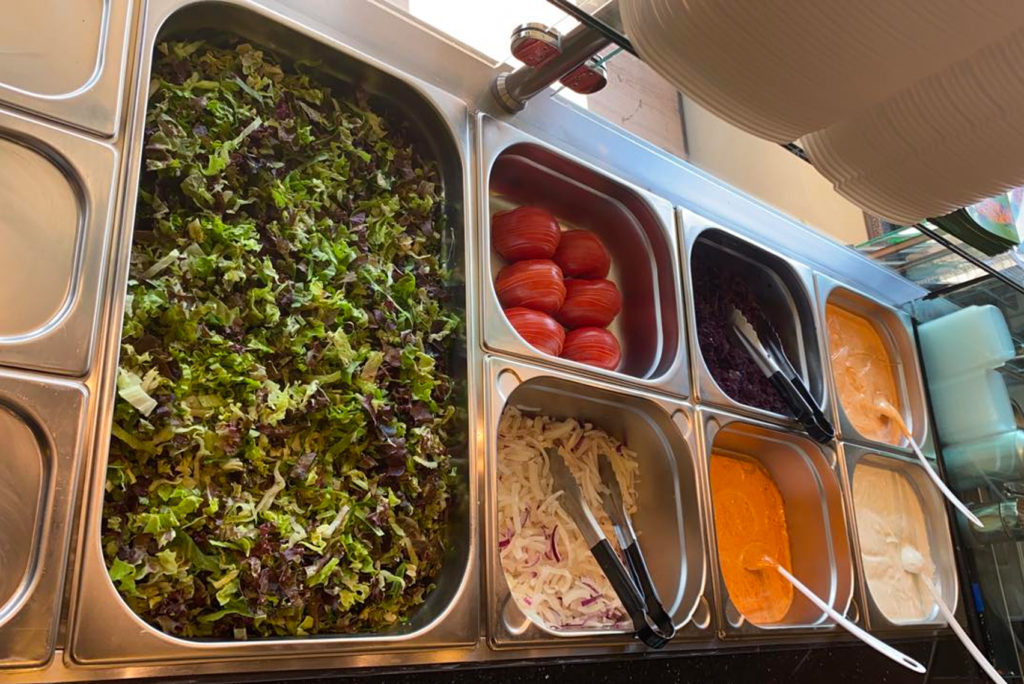 Eine Salattheke mit Tabletts mit gemischtem Blattgemüse, geschnittenen Tomaten, zerkleinertem Kohl, Rotkohl und Behältern mit orangefarbenem und weißem Dressing. Zum Servieren liegen Zangen und Löffel in den Tabletts.