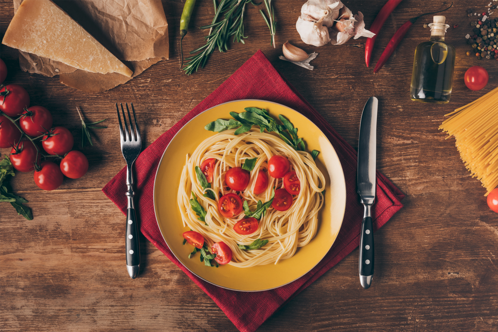 Ein Teller Spaghetti mit Kirschtomaten und Gemüse auf einem gelben Teller, auf einer roten Serviette. Umgeben von Parmesan, Kirschtomaten, Knoblauch, Kräutern, Chilischoten, Olivenöl und ungekochten Spaghetti auf einem Holztisch.