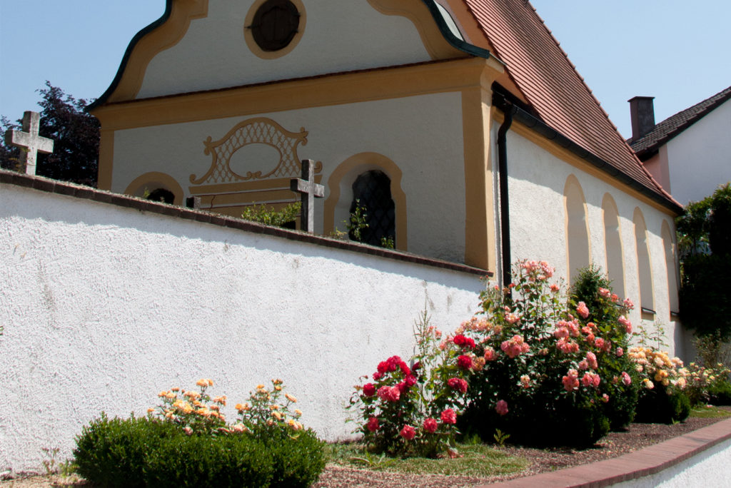 Eine kleine Kapelle mit einem braunen Ziegeldach und einer weißen Außenfassade mit gelben Verzierungen. Davor säumt ein Garten mit leuchtend roten, rosa und gelben Blumen die Basis einer weißen Mauer. Auf der linken Seite der Mauer sind zwei Steinkreuze zu sehen. Der Himmel ist klar und blau.