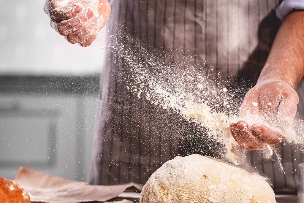 Eine Person mit Schürze streut Mehl über einen Teigballen auf einer Küchentheke. Die Szene fängt die Bewegung des Mehlstaubs in der Luft ein und betont den Backvorgang.