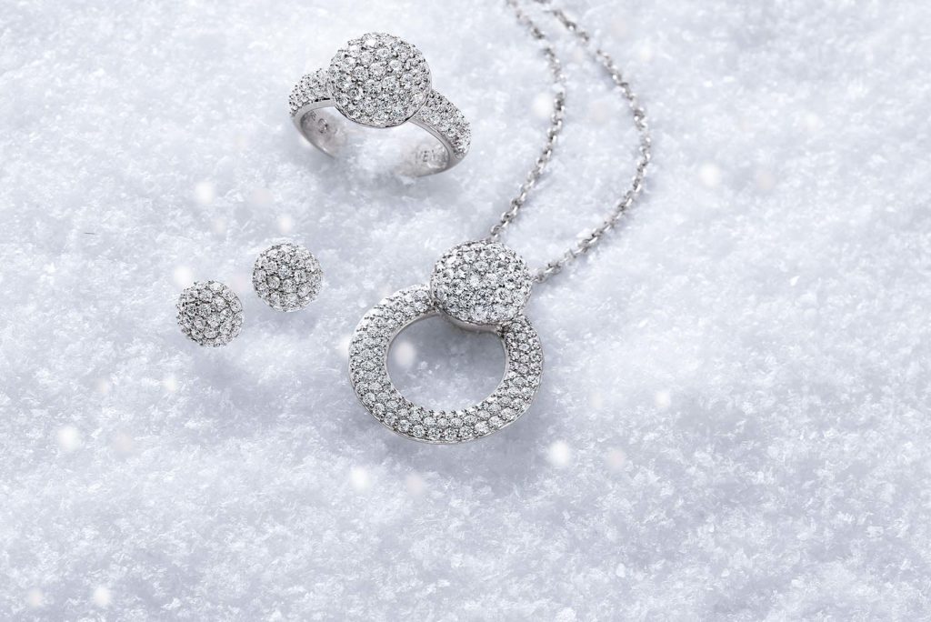 Ein luxuriöses Schmuckset bestehend aus einem diamantbesetzten Ring, Ohrringen und einer Halskette mit rundem Anhänger, alles präsentiert auf einem weichen, weißen Schneehintergrund.