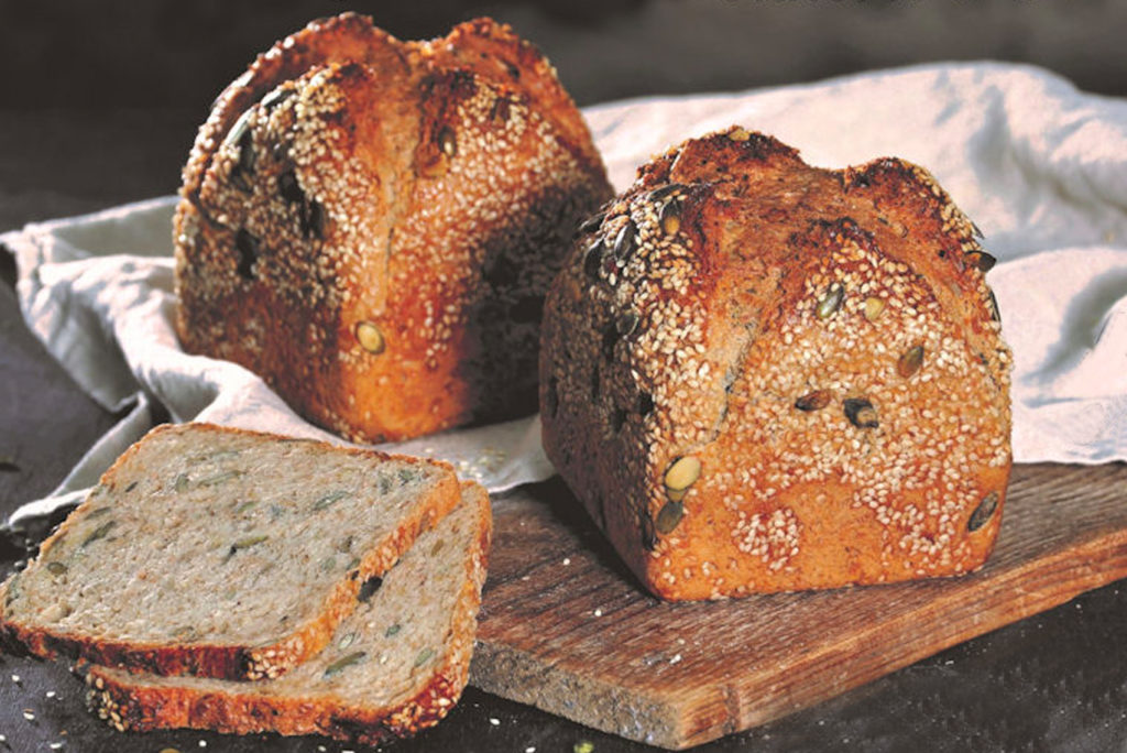 Zwei Laibe Mehrkornbrot, belegt mit Körnern, möglicherweise Sesam und Kürbis, liegen auf einem Holzbrett. Zwei Scheiben sind abgeschnitten und daneben auf ein weißes Tuch gelegt. Das Brot wirkt herzhaft und strukturiert.