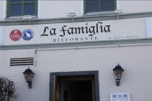 Der Eingang eines Restaurants mit einem Schild mit der Aufschrift „La famiglia Ristorante da Renato Bordini“, zwei Logos auf der linken Seite und zwei Wandlaternen auf beiden Seiten des Eingangs.