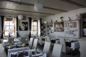 Ein helles, modernes Restaurant mit weißen Stühlen und braunen Tischdecken. Die Wände sind mit gerahmten Fotos, Gemälden und Topfpflanzen dekoriert. Große Fenster lassen viel Tageslicht herein.