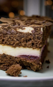 Eine Nahaufnahme eines geschichteten Schokoladenkuchenstücks mit einer cremeweißen Füllung, einer dünnen Schicht Fruchtmarmelade und Schokoladenraspeln obenauf.