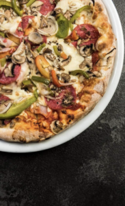 Eine Nahaufnahme einer Pizza auf einem weißen Teller, belegt mit Pilzen, grünen und gelben Paprikaschoten und Peperonischeiben, auf einer dunklen strukturierten Oberfläche.