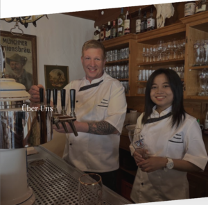 Zwei lächelnde Mitarbeiter in weißen Uniformen stehen hinter einer Bar; einer gießt ein Getränk aus einem Hahn ein, während der andere ein Glas hält. Die Bar ist mit Gläsern, Flaschen und Holzakzenten dekoriert. Der Schriftzug "Über Uns" erscheint auf dem Bild.