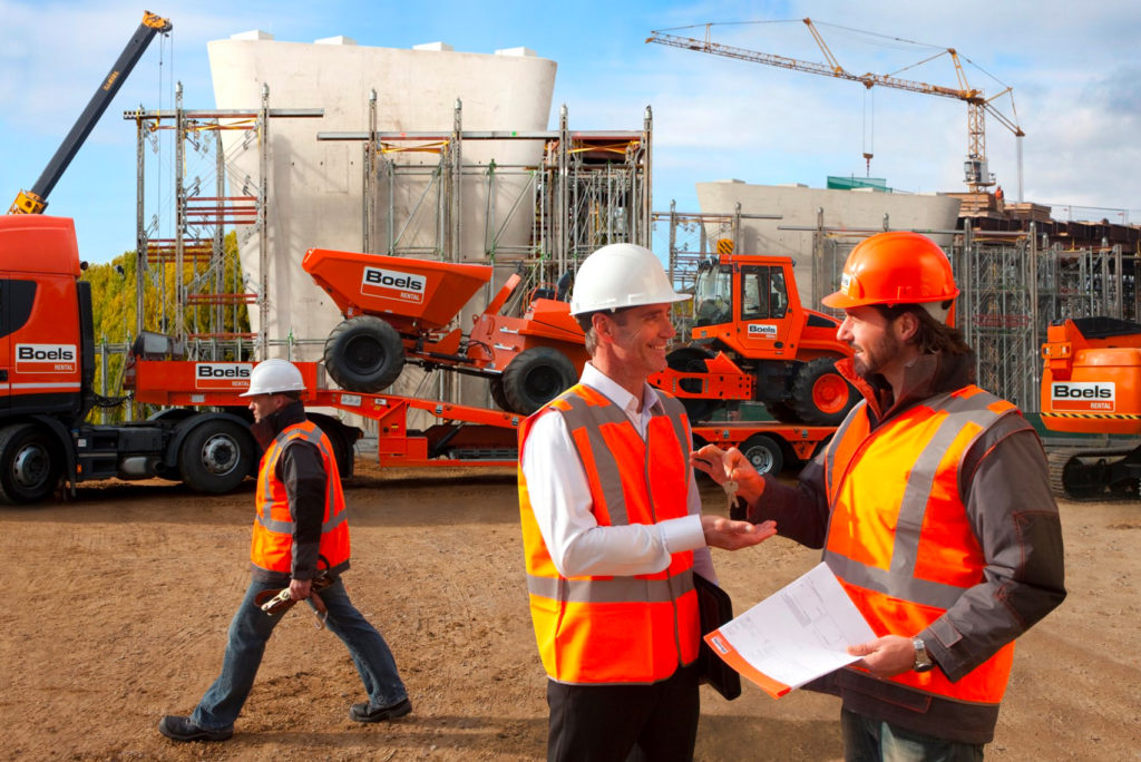 Drei Bauarbeiter in Schutzhelmen und orangefarbenen Westen sind auf einer Baustelle. Zwei schütteln sich die Hände und unterhalten sich, einer hält Papiere in der Hand. Auf der Baustelle gibt es Gerüste, Fahrzeuge und Kräne. Im Hintergrund ist eine große Betonkonstruktion zu sehen.