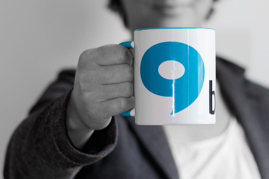 Eine Person im Anzug hält einen weißen Becher mit der großen blauen Zahl Neun darauf. Der Hintergrund ist in Graustufen verschwommen und konzentriert sich auf den Becher und einen Teil der Hand und Kleidung der Person.
