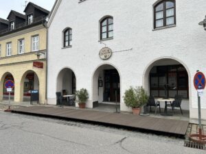 In einer ruhigen Straße liegt ein Café mit gewölbten Eingängen und Sitzgelegenheiten im Freien auf einer Holzplattform. Zwei Parkverbotsschilder sind sichtbar, und neben den Tischen stehen Topfpflanzen. Das Gebäude hat weiße Wände und Fenster im Obergeschoss.