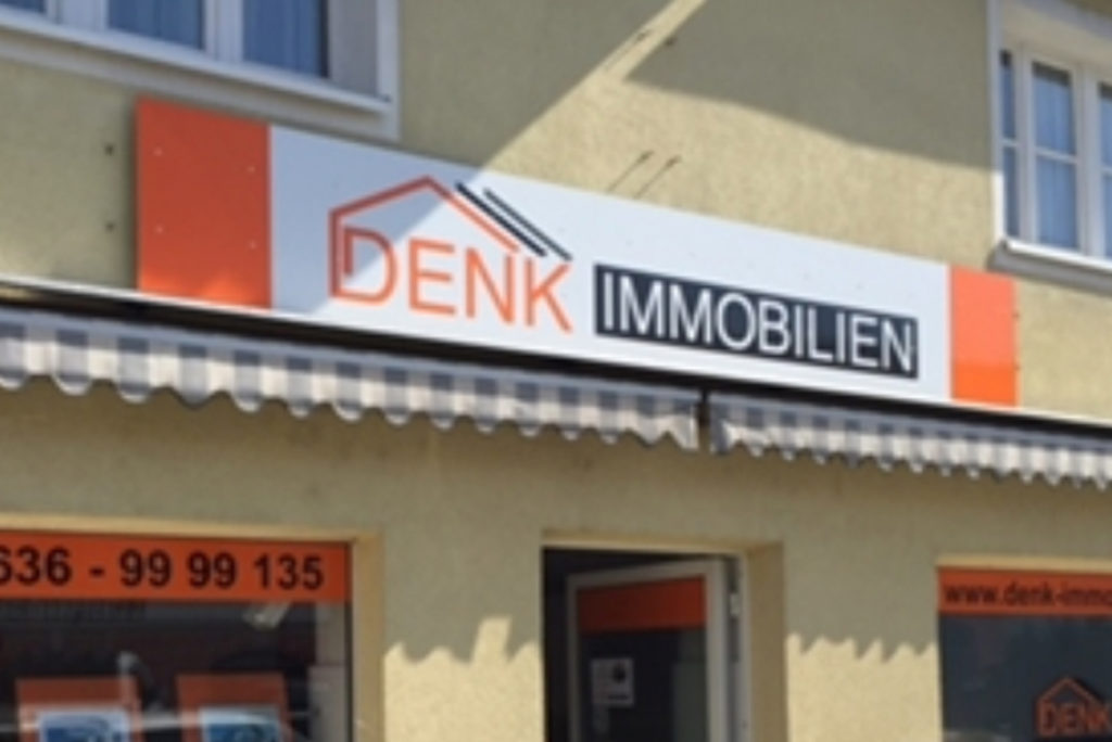 Das Bild zeigt eine Ladenfront mit einem Schild mit der Aufschrift „DENK IMMOBILIEN“. Das Schild zeigt die Grafik eines Hausdachs über dem Wort „DENK“. Die Ladenfront ist gelb gestrichen, hat eine Markise und in einem Fenster ist die Kontaktnummer des Unternehmens zu sehen.