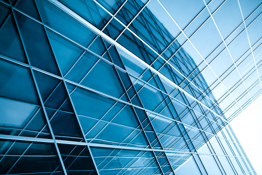 Nahaufnahme einer modernen Gebäudefassade mit reflektierenden Glasplatten und einem Gittermuster. Das Glas reflektiert einen blauen Himmel und erzeugt ein dynamisches und abstraktes architektonisches Design mit sich kreuzenden Linien und Winkeln.