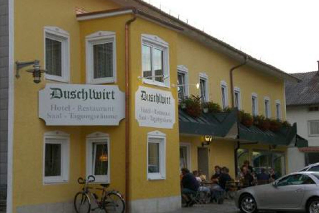 Ein gelbes Gebäude mit Schildern mit der Aufschrift „Hotel - Restaurant“ und mehreren Fenstern mit Blumenkästen. Menschen sitzen draußen unter grünen Markisen, an der Wand steht ein Fahrrad. In der Nähe parkt ein Auto.