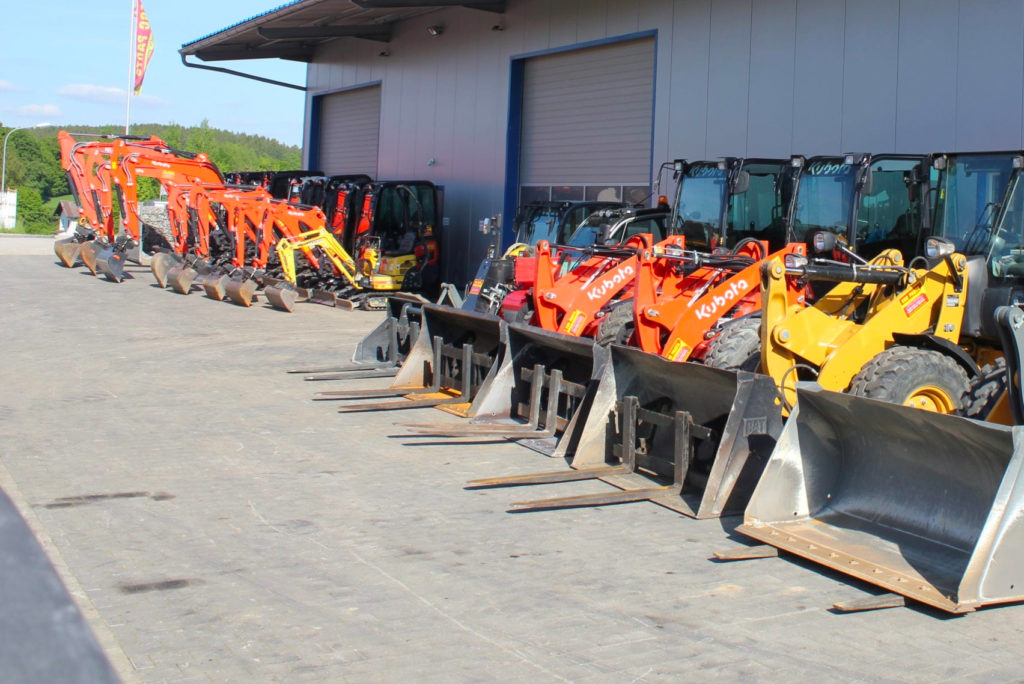 Reihen roter und gelber Baufahrzeuge, darunter Lader und Bagger, stehen auf einem gepflasterten Außengelände neben einem großen Industriegebäude. Der Himmel ist klar und die Umgebung ist von grünen Bäumen gesäumt.
