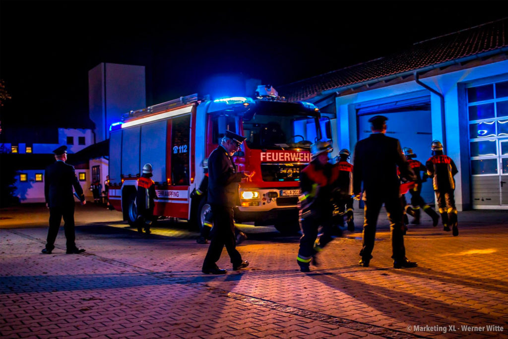 Feuerwehrleute in Uniform stehen nachts neben einem hell erleuchteten Feuerwehrauto mit der Aufschrift „FEUERWEHR“ vor einem Gebäude und bewegen sich dort. Blaue Lichter am Auto erhellen die Szene, während sich das Team auf den Einsatz vorbereitet.