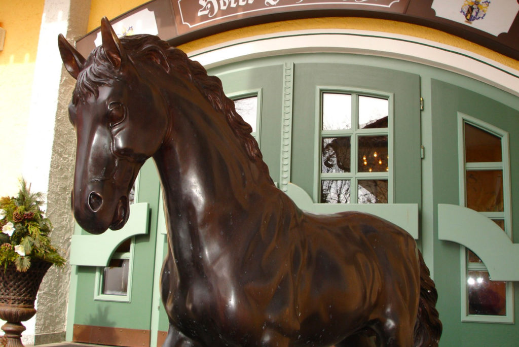 Eine dunkelbraune Pferdestatue steht vor einem Gebäude mit kunstvollen grünen und weißen Türen. Links sind dekorative Pflanzen zu sehen. Das Gebäude hat gelbe Wände und über den Türen ein Schild mit dekorativen Mustern.