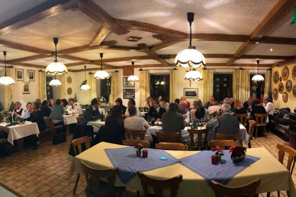 Ein warm beleuchtetes Restaurant mit Holzbalken und -wänden, in dem eine große Gruppe von Menschen an Tischen mit weißen Tischdecken und blauen Läufern sitzt. Die Tische sind mit kleinen Blumenarrangements und Kerzen dekoriert, was eine gemütliche Atmosphäre schafft.