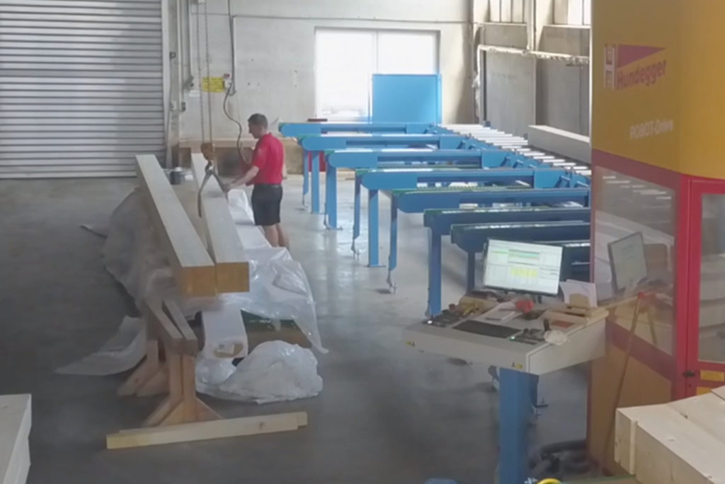 Ein Arbeiter im roten Hemd bedient Maschinen in einer großen Industrieanlage. Auf einem Tisch neben ihm liegen lange Balken, und ein Computerbildschirm zeigt Daten an. Durch ein Fenster im Hintergrund fällt helles Licht ein.