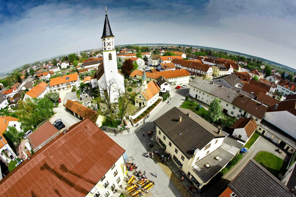 Luftaufnahme eines kleinen Dorfs mit einer markanten weißen Kirche und einem hohen Kirchturm, wo der Tag damit beginnt, dass sich die Menschen auf einem Freiluftmarkt versammeln. Die umliegenden Gebäude mit roten und braunen Dächern verleihen der Szenerie Charme, während von Bäumen gesäumte Straßen zum Erkunden einladen.