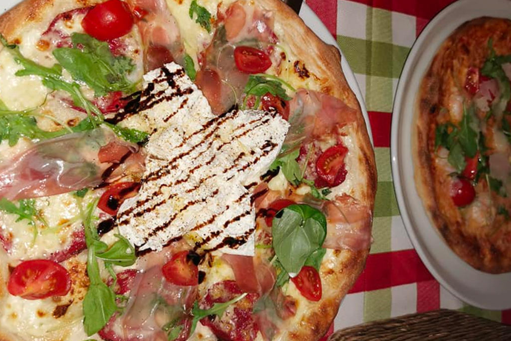 Pizza mit Rucola, Kirschtomaten, Wurst und Ricotta-Käse, beträufelt mit Balsamico-Glasur, serviert auf einem Teller. Eine andere, teilweise sichtbare, ähnliche Pizza liegt daneben auf einer karierten Tischdecke.