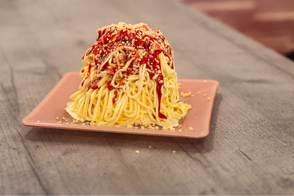Ein Dessert, das an Spaghetti erinnert, wird auf einem rosafarbenen, quadratischen Teller serviert. Es besteht aus Vanilleeis in Nudelform, garniert mit Erdbeersauce im Marinara-Stil und bestreut mit weißer Schokolade, die an Parmesankäse erinnert.