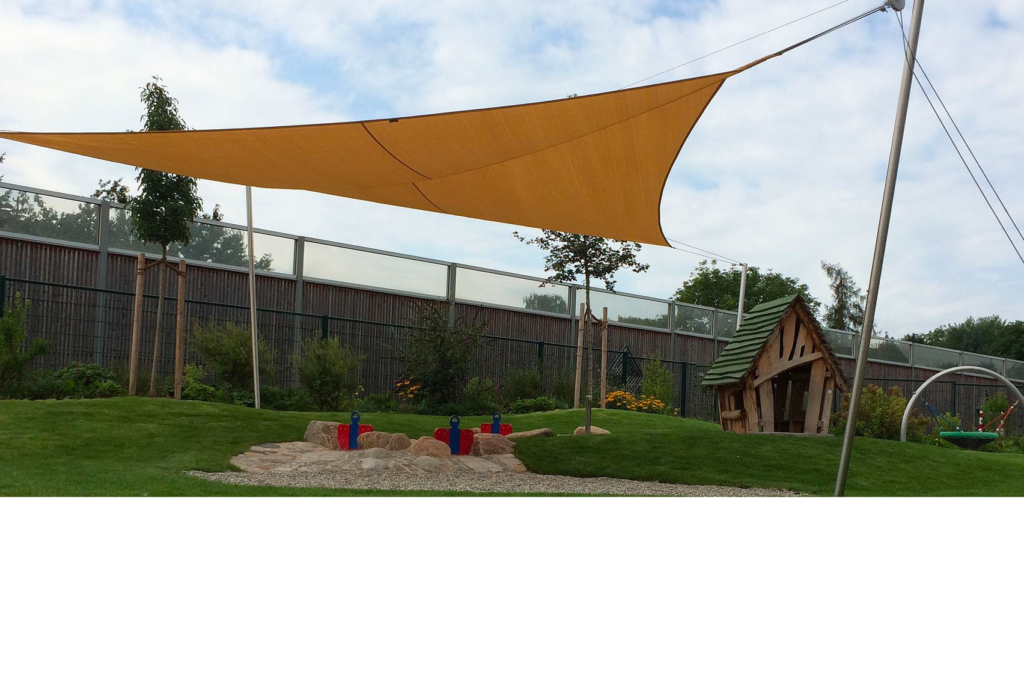 Ein Spielplatz mit einem schattigen Sandkasten unter einem großen dreieckigen Baldachin. Um den Sandkasten herum stehen kleine Bäume und Sträucher. Im Hintergrund ist ein kleines Holzspielhaus zu sehen. Der Himmel ist teilweise bewölkt.