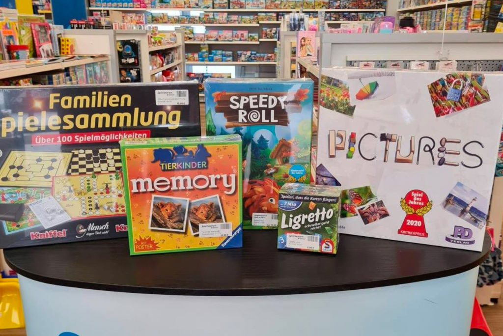 Brettspiele auf einem Tisch in einem Geschäft. Zu den Spielen gehören „Familienspielesammlung“, „Speedy Roll“, „Pictures“, „Tierkinder Memory“ und „Ligretto“, umgeben von Regalen voller verschiedener Spielsachen und Spiele. Helle und farbenfrohe Umgebung.
