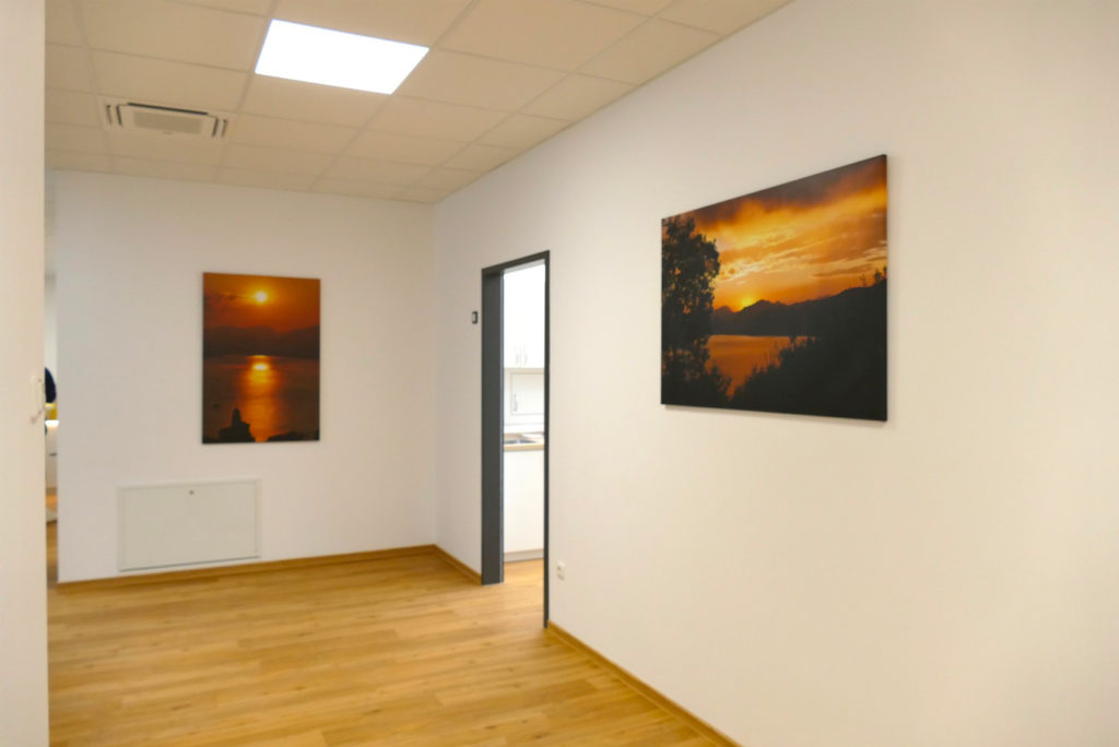 In einem Flur mit Holzfußboden und weißen Wänden hängen zwei Fotos von Sonnenuntergängen. Ein Foto hängt an der linken Wand, das andere an der gegenüberliegenden. Eine offene Tür führt in einen anderen Raum, in dem man ein Waschbecken erspäht. An der Decke hängen Leuchtstoffröhren.