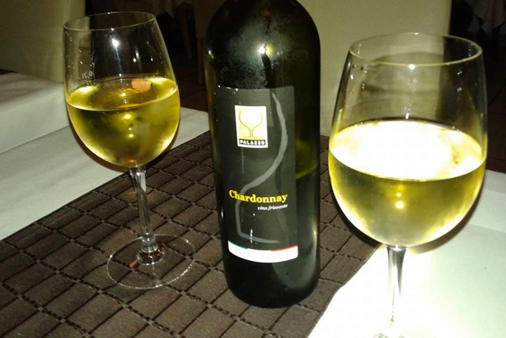 Eine Flasche Chardonnay-Wein steht auf einer dunklen gewebten Matte, daneben stehen zwei mit Weißwein gefüllte Gläser auf einem mit einer weißen Tischdecke gedeckten Tisch.