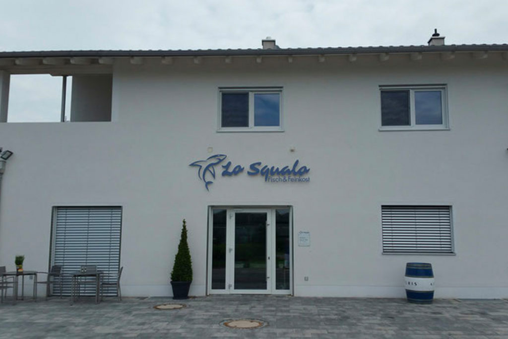 Weißes, zweistöckiges Gebäude mit einem Schild mit der Aufschrift „Lo Squalo“ neben einem Hai-Logo. Die Fassade hat Fenster mit Fensterläden und ein Fass neben einer Tür. Draußen stehen Tische und Stühle auf dem Steinpflaster.