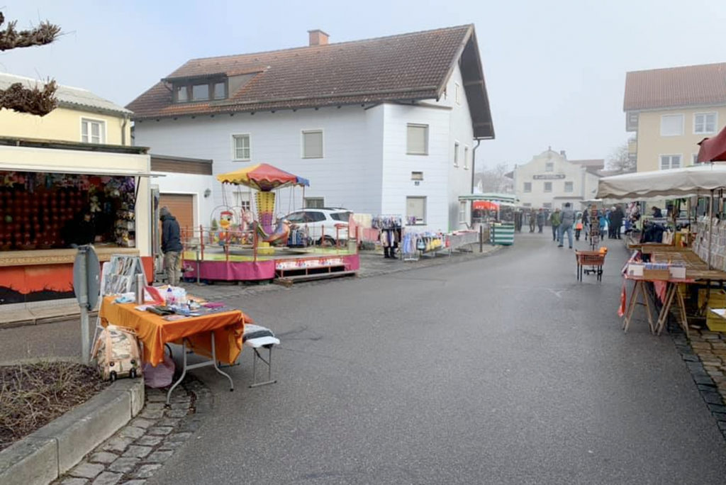 Ein kleiner Straßenmarkt mit verschiedenen Ständen, an denen Waren wie Kleidung und Kunsthandwerk verkauft werden. Es gibt einen Imbisswagen und eine Auswahl an Artikeln auf Tischen. Das Gebiet ist gesäumt von Wohnhäusern unter einem bewölkten Himmel. Es sind nur wenige Menschen unterwegs.