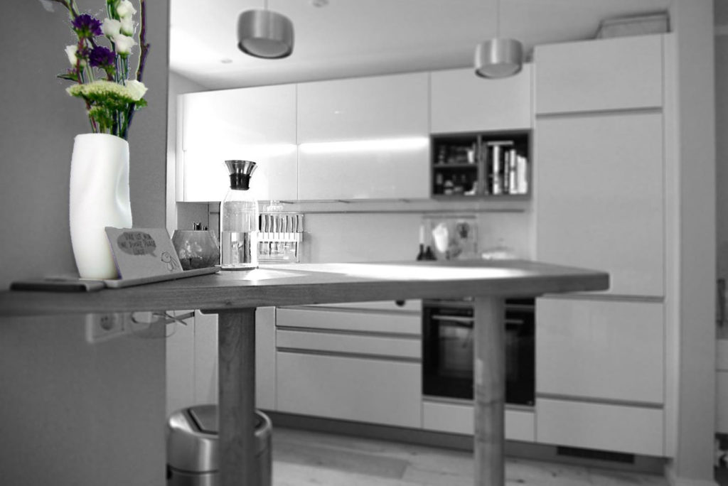 Moderne Küche in Schwarzweiß mit einer hohen, weißen Vase mit violetten und weißen Blumen auf einer Holztheke. Im Hintergrund sind helle Schränke mit Regalen, ein Ofen und andere Küchenutensilien zu sehen.