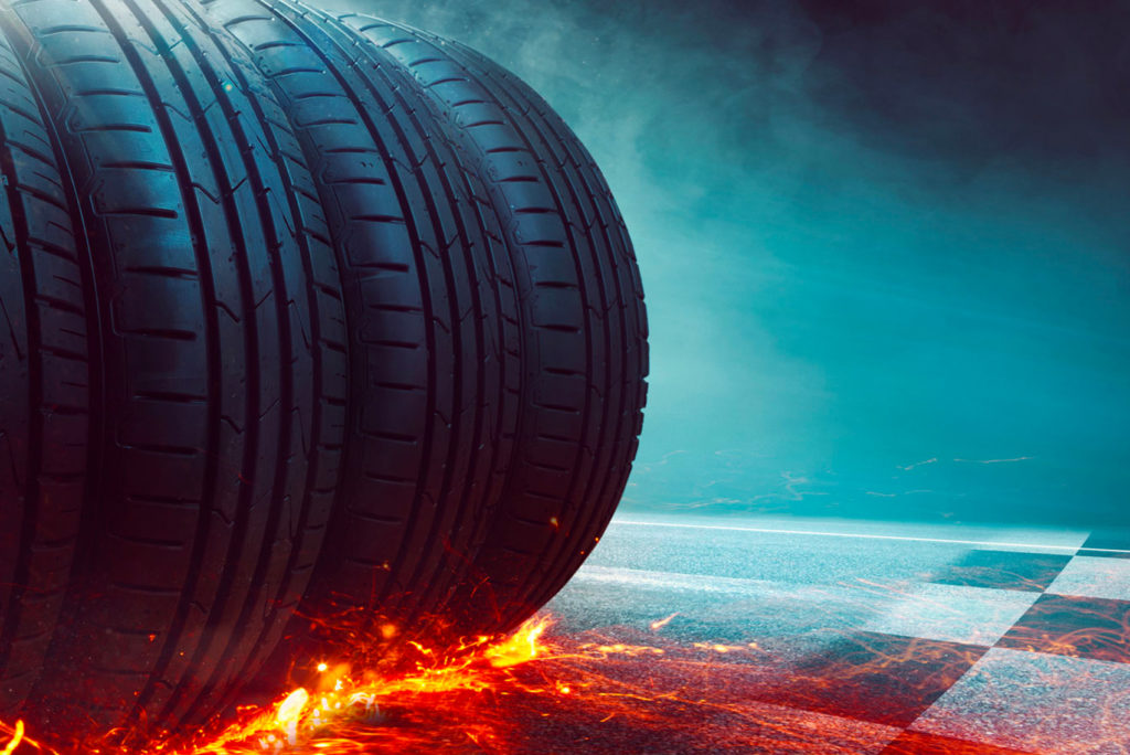 Ein Satz Autoreifen auf einer Straße mit roten Flammen darunter, die einen Eindruck von Geschwindigkeit und Intensität vermitteln. Der Hintergrund ist ein bläulicher Himmel mit einem Hauch von Wolken.