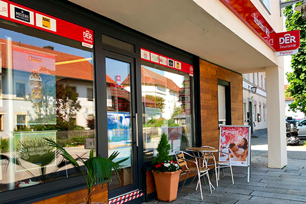Außenansicht eines Reisebüros mit einem großen Fenster, in dem Werbeplakate hängen. Auf dem Gehsteig steht ein kleiner Tisch mit zwei Stühlen und einer Topfpflanze. Auf dem Schild steht „DER Touristik“. In dem Fenster spiegeln sich nahe gelegene Gebäude und ein Baum.
