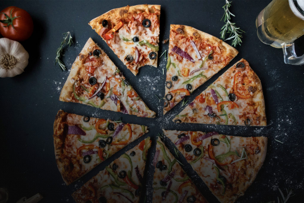 Eine Pizza in Scheiben mit verschiedenen Belägen, darunter Oliven, Paprika und Zwiebeln, auf einer dunklen Oberfläche. Aus einem Stück ist ein Stück herausgeschnitten. Daneben liegen eine Tomate, Knoblauch, ein Zweig Rosmarin und ein Krug Bier. Auf der Oberfläche sind Mehlstreusel zu sehen.