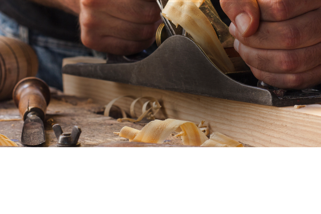 Hände, die mit einem Hobel ein Holzbrett hobeln, umgeben von Holzspänen und verschiedenen Schnitzwerkzeugen.
