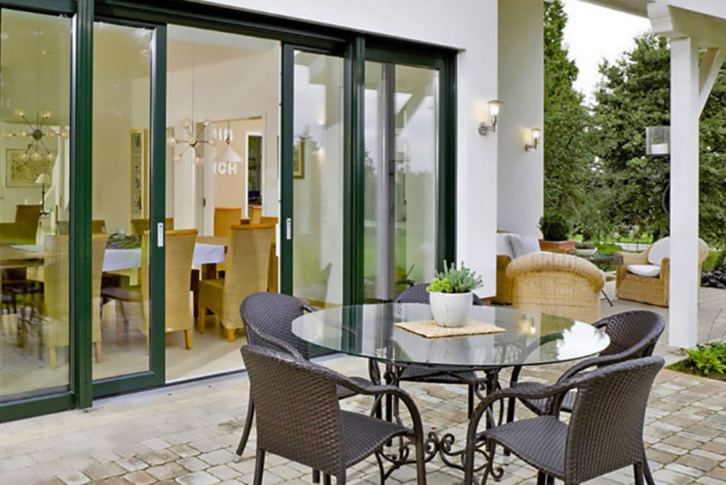 Eine Terrasse mit einem runden Glastisch und vier Korbstühlen. Glasschiebetüren führen zu einem Essbereich im Innenbereich. Auf der rechten Seite befinden sich Sitzgelegenheiten im Freien mit einem Sofa und einem Sessel, umgeben von Grün und einem gepflasterten Steinboden.