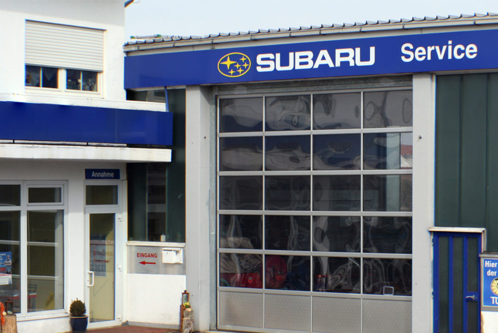 Subaru-Servicecenter mit blauem Schild und Logo auf einem großen Garagentor. Links ist ein kleiner Eingang mit Glastür, beschriftet mit „Annahme“ und „Eingang“. Das Gebäude hat weiße und grüne Wände.