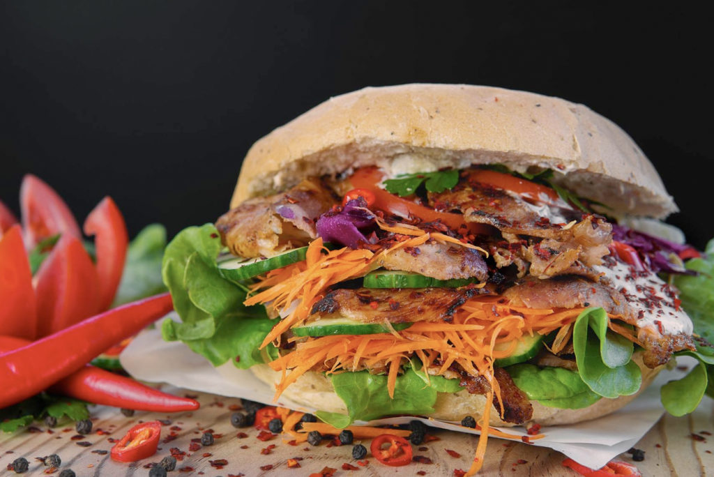 Ein herzhaftes Sandwich mit gegrilltem Fleisch, geraspelten Karotten, Salat, Gurke und Tomatenscheiben zwischen einem knusprigen Brötchen. Dazu gibt es rote Chilischoten und Tomatenscheiben. Garniert wird das Sandwich mit Kräutern und Chiliflocken.