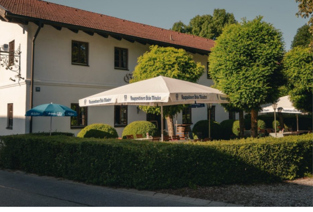 Ein weißes Gebäude mit rotem Ziegeldach bietet an sonnigen Tagen einen schattigen Sitzplatz im Freien mit großen weißen und blauen Sonnenschirmen, umgeben von ordentlich gestutzten Hecken und Bäumen.