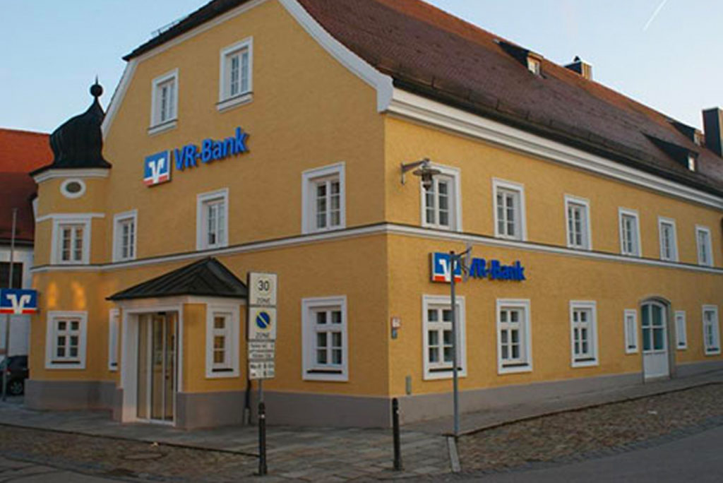Ein gelbes Gebäude mit rotem Dach beherbergt eine Filiale der VR-Bank. Das Gebäude verfügt über mehrere Fenster und ein kleines Eingangsvordach. Davor ist ein Straßenschild zu sehen, das Tempo 30 und eine Parkverbotszone anzeigt. Der Himmel ist klar.