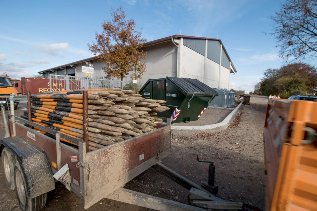 Eine Baustelle mit einem Gebäude im Hintergrund. Im Vordergrund steht ein mit Holzpfosten beladener Anhänger, daneben steht ein grüner Müllcontainer. Blattlose Bäume flankieren die Baustelle unter einem teilweise bewölkten Himmel.