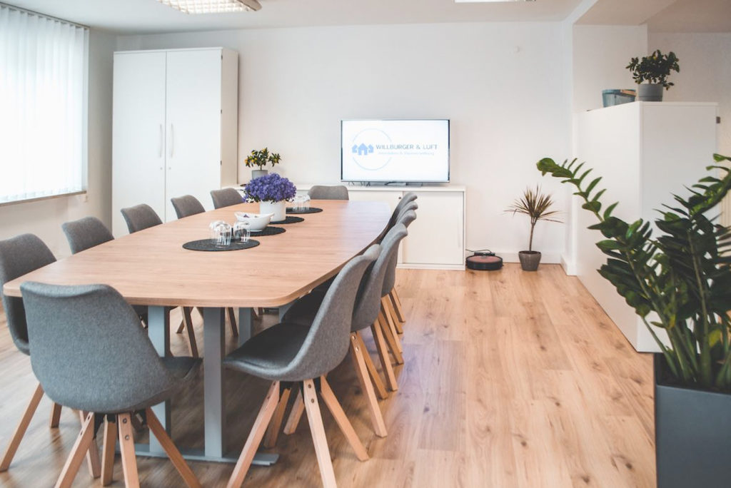 Ein moderner Konferenzraum mit einem langen Holztisch, umgeben von grauen Stühlen. An der Wand ist ein Fernsehbildschirm mit einem Logo angebracht. Topfpflanzen und ein Schrank sind vorhanden. Der Raum hat Holzboden und weiße Wände.