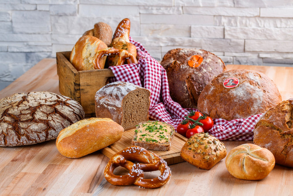 Verschiedene Brotsorten und Brezeln, darunter Laibe, Brötchen und geschnittenes Brot, sind auf einem Holztisch mit einer rotkarierten Tischdecke arrangiert. Daneben steht ein Bündel Kirschtomaten, die für einen Hauch Farbe sorgen. Eine rustikale Steinmauer bildet den Hintergrund.
