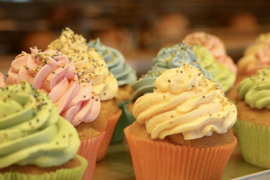 Bunte Cupcakes mit gewirbeltem Zuckerguss und Streuseln auf einem Tablett. Die Farben des Zuckergusses sind Rosa, Grün, Blau und Gelb. Die Cupcake-Verpackungen sind orange und gelb. Der Hintergrund ist unscharf und konzentriert sich auf die Cupcakes.