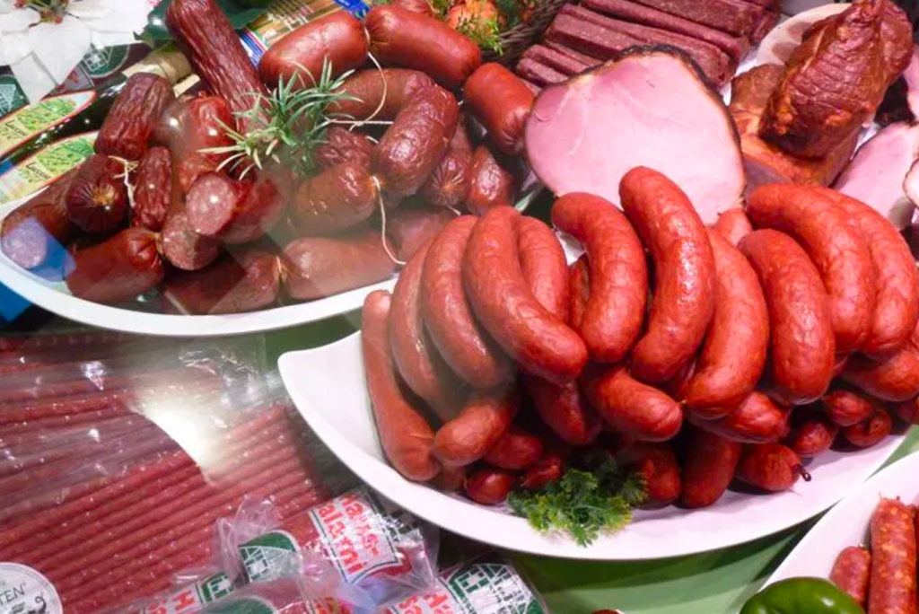 Eine Auswahl verschiedener Wurstwaren, darunter Stapel von Würsten und Schinkenscheiben auf weißen Tellern. Die Wurstwaren sind von verpackten Würsten umgeben, und ein Zweig Thymian verleiht ihnen einen Hauch von Grün.