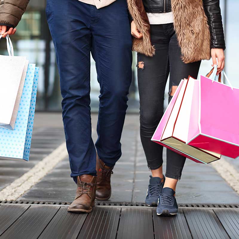 Ein Paar geht zusammen mit Einkaufstüten. Die Person links trägt blaue Hosen und braune Schuhe, während die Person rechts zerrissene Jeans, eine Pelzweste und Turnschuhe trägt. Sie stehen auf einem Bürgersteig in der Stadt.