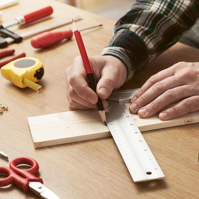 Eine Person in einem karierten Hemd verwendet ein Lineal und einen Bleistift, um ein Holzbrett auf einer Werkbank zu messen und zu markieren. Um sie herum liegen verschiedene Werkzeuge wie ein Maßband, Schraubendreher und Scheren.