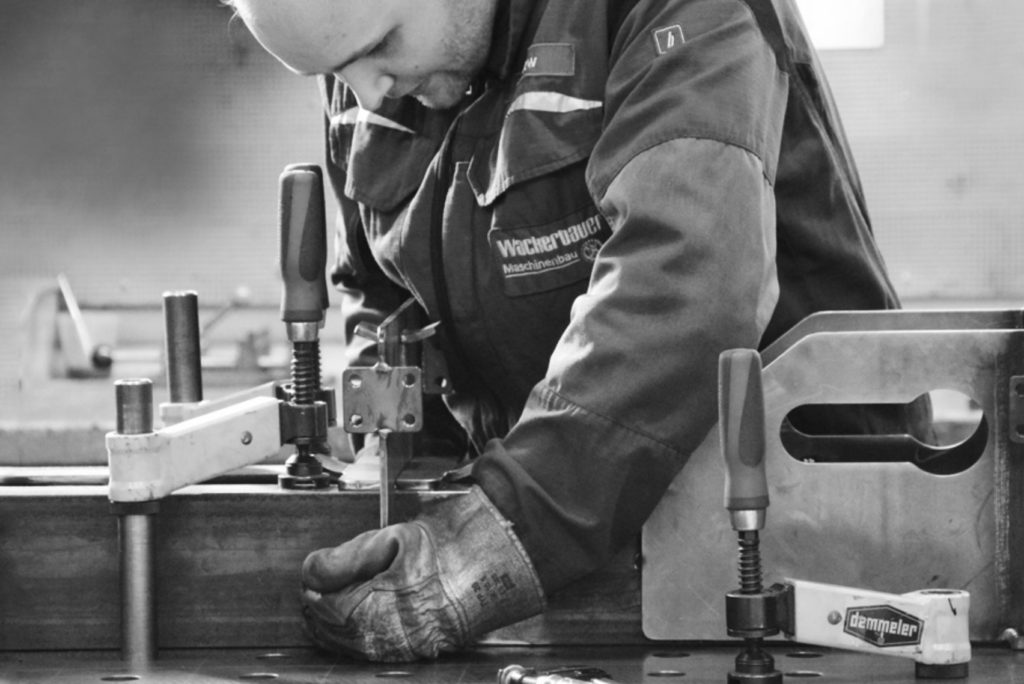 Eine Person mit Schutzhandschuhen und Overall schweißt oder bearbeitet ein Metallstück und hält die Teile mithilfe von Klammern auf einer Werkbank fest. Die Szene ist in Schwarzweiß gehalten und zeigt eine konzentrierte Industrie- oder Werkstattumgebung.